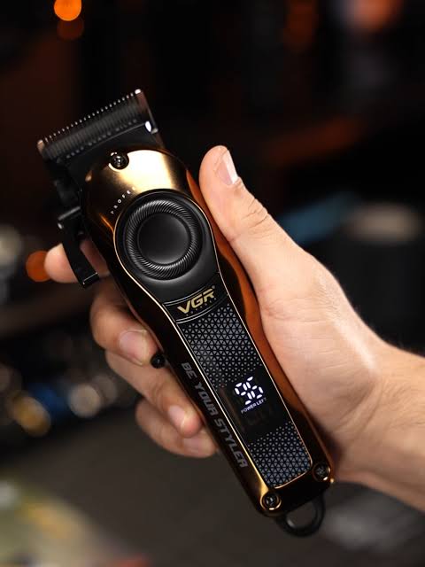 A close-up of a hand holding a metallic gold and black VGR electric hair clipper against a dark, blurred background.